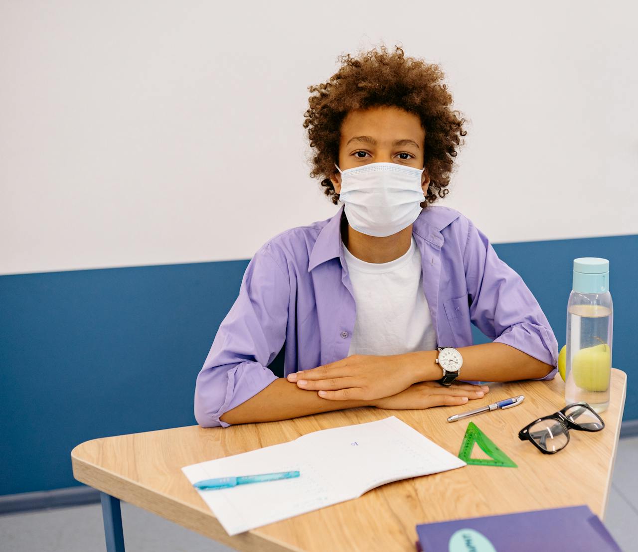 Young student wearing face mask sitting at school desk with supplies, emphasizing new normal education.